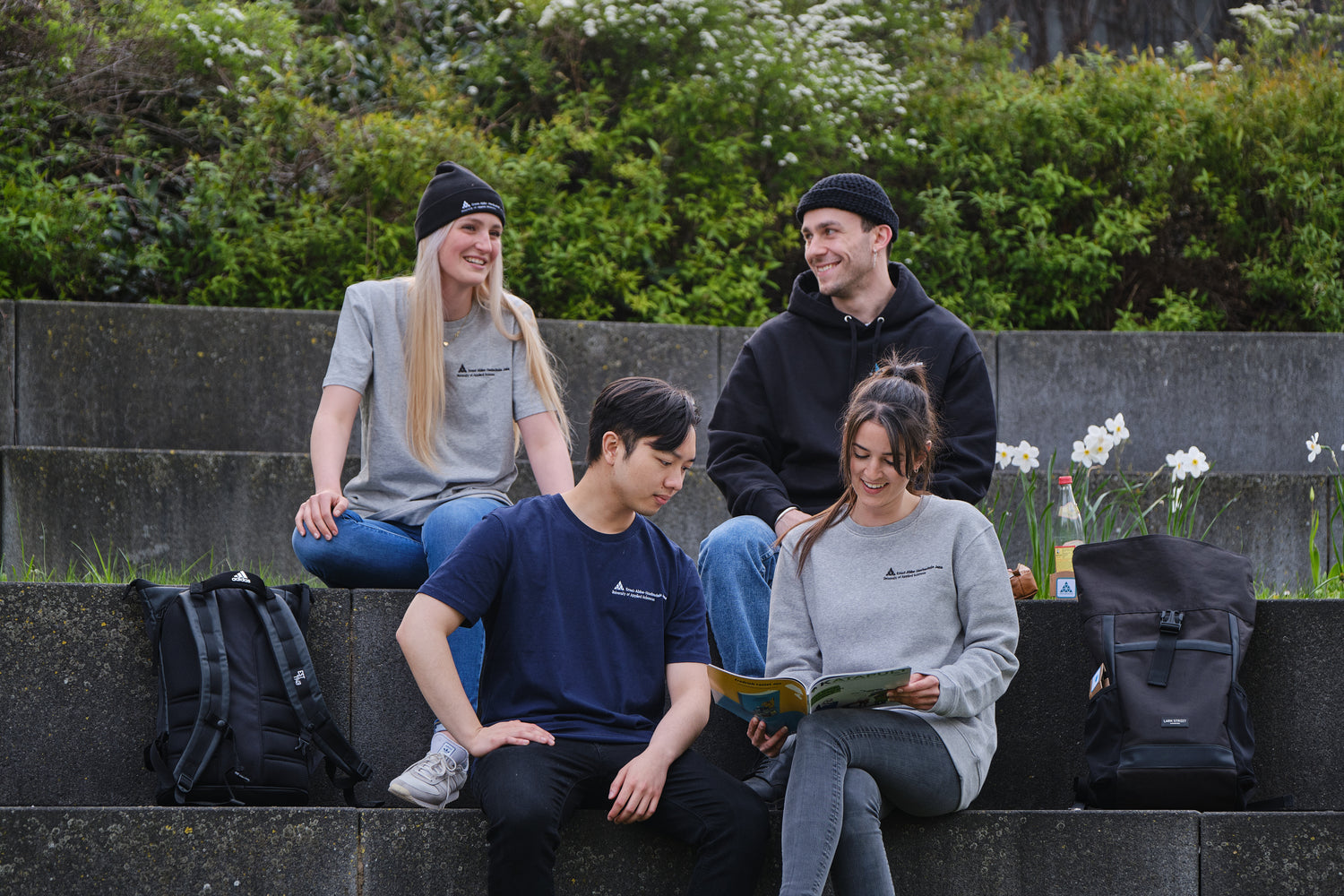 2 Studenten und 2 Studentinnen sitzen auf Steinstufen und tragen Kleidung von StudyMerch, die mit dem Logo der Ernst-Abbe-Hochschule veredelt ist. Zu sehen sind ein graues und ein dunkelblaues T-Shirt, ein grauer Sweater und ein schwarzer Hoodie.