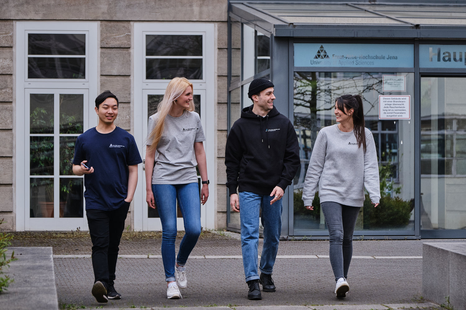 2 Studenten und 2 Studentinnen laufen auf die Kamera zu und unterhalten sich. Sie tragen Kleidung von StudyMerch, die mit dem Logo der Ernst-Abbe-Hochschule veredelt ist: v.l.n.r. dunkelblaues und graues T-Shirt, schwarzer Hoodie und grauer Sweater.