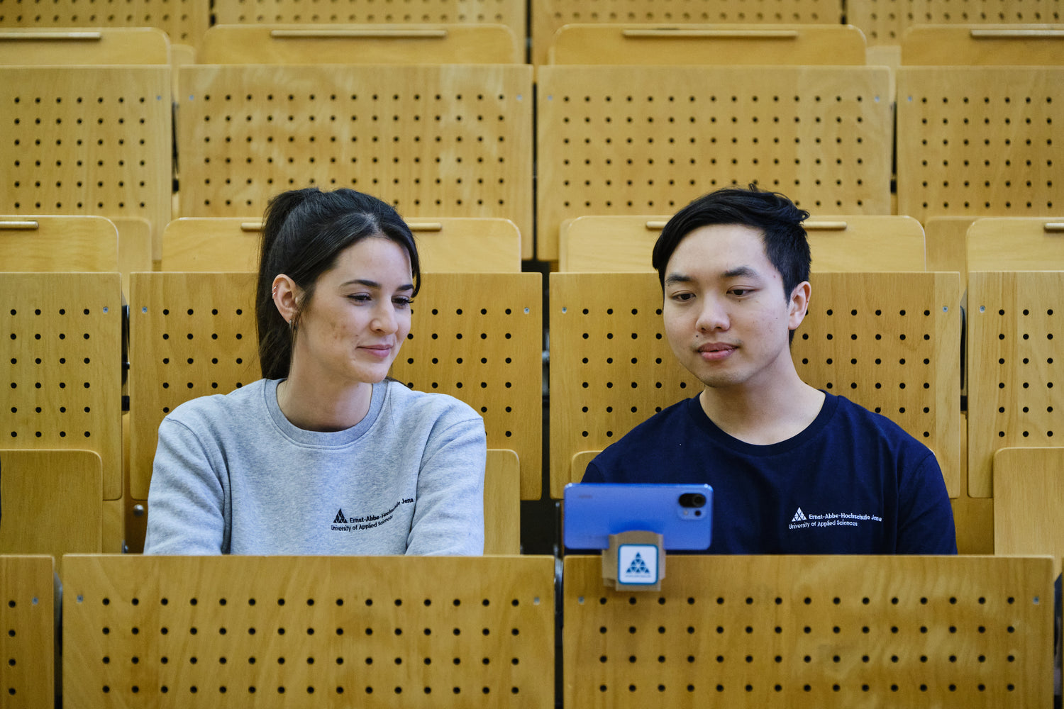 Ein Student und eine Studentin sitzen im Hörsaal und tragen einen grauen Sweater und ein dunkelblaues T-Shirts von StudyMerch, die mit dem Logo der Ernst-Abbe-Hochschule Jena veredelt sind. Vor den Studierenden befindet sich der StudyBuddy, ein Gadget in welchem hier ein Handy abgestellt wurde.