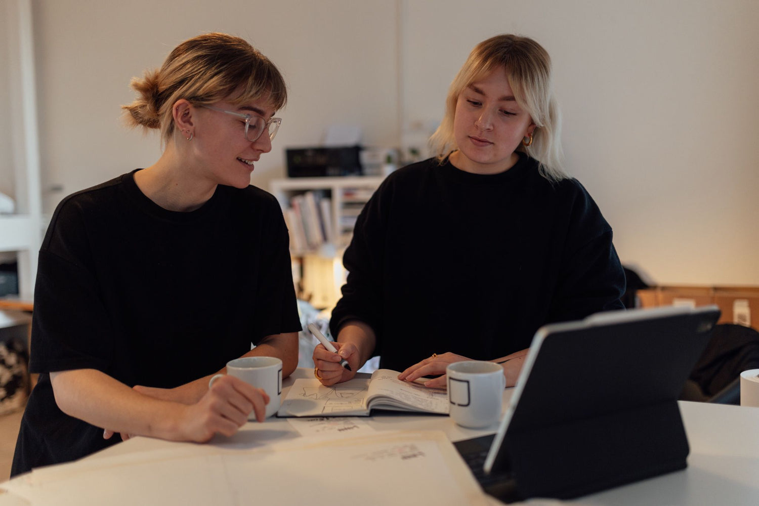 Designer-Duo Lex & Julez steht beim Designen ihrer nachhaltigen Mode mit einem iPad, 2 Kaffeetassen und einem Notizbuch an einem Tisch