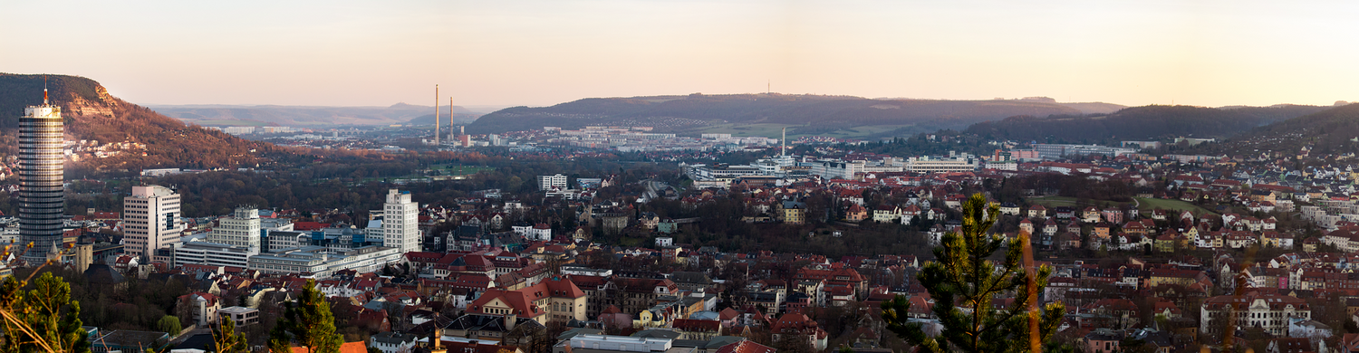 Blick auf die Stadt Jena mit den angrenzenden Hügeln im Hintergrund und dem JenTower am linken Bildrand